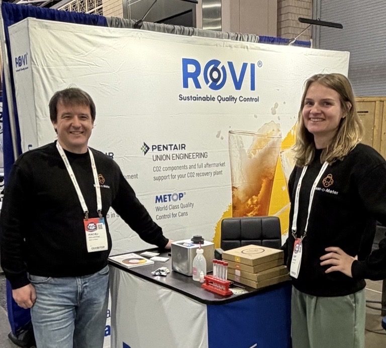 Two people standing at a ROVI trade show booth, presenting quality control equipment and demonstration materials on a counter, with a branded company banner in the background.