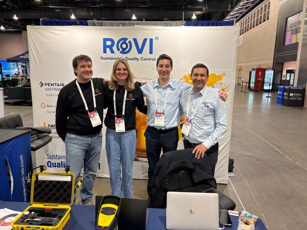 People standing at a ROVI trade show booth, presenting quality control equipment and demonstration materials on a counter, with a branded company banner in the background.